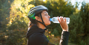 Mujer con casco de bicicleta y ropa deportiva bebiendo de una botella de agua con árboles a su alrededor