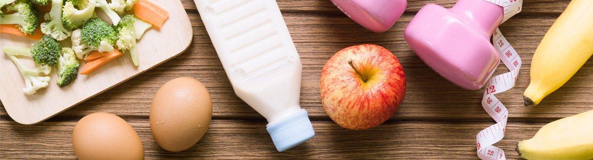 Mesa de elementos de un estilo de vida saludable, como: mancuerdas, metro, una bebida y alimentos saludables como: frutas, verduras, huevos.