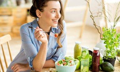 Mujer sonriente apoya su brazo en una mesa mientras come frutas frescas.