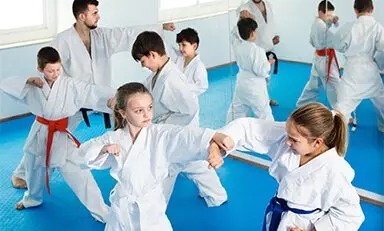 Grupo de personas practicando artes marciales en un dojo, vestidos con uniformes tradicionales blancos llamados gi y cintos de colores que indican su rango. Están realizando ejercicios de entrenamiento, como golpes y posturas, en una sala con suelo de tatami y espejos en la pared