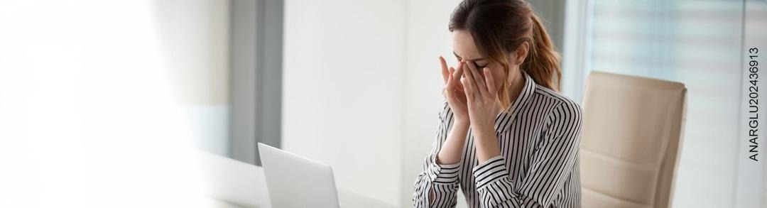 Mujer sosteniendo sus sienes en señal de dolor, sentada en una oficina frente a su computador
