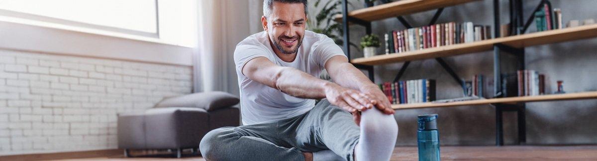  Hombre sonriente sentado en el piso, junto a un estante con libros, se toca la punta del pie
