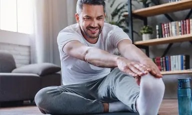  Hombre sonriente sentado en el piso, junto a un estante con libros, se toca la punta del pie