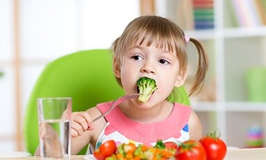 un niño sentado a la mesa con un plato de comida y un vaso de agua. El niño sostiene un tenedor y parece estar a punto de comer. El fondo incluye una silla verde y una estantería con varios objetos