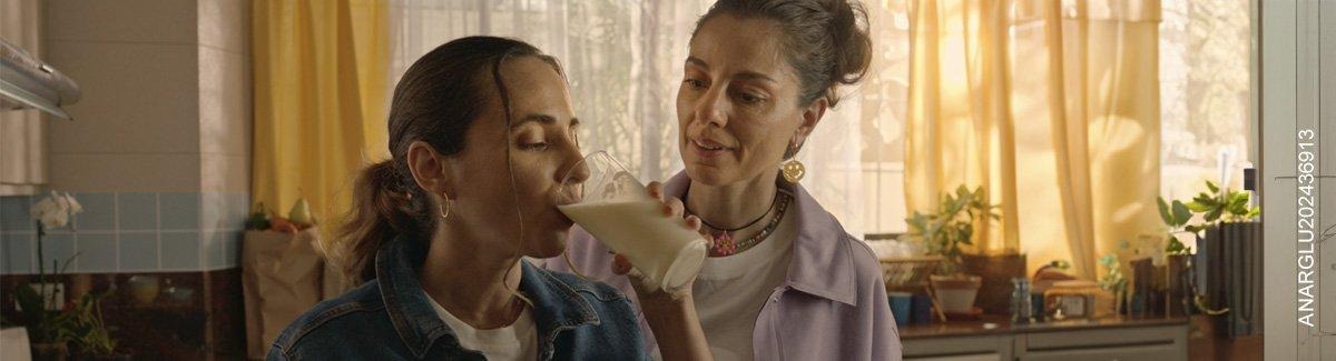 Mujeres en la cocina, una está tomando una bebida y la otra la está observando con gesto de aprobación.
