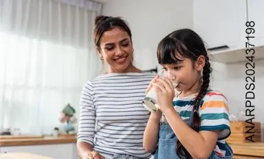 Niña toma de un vaso mientras su mamá la mira sonriendo.