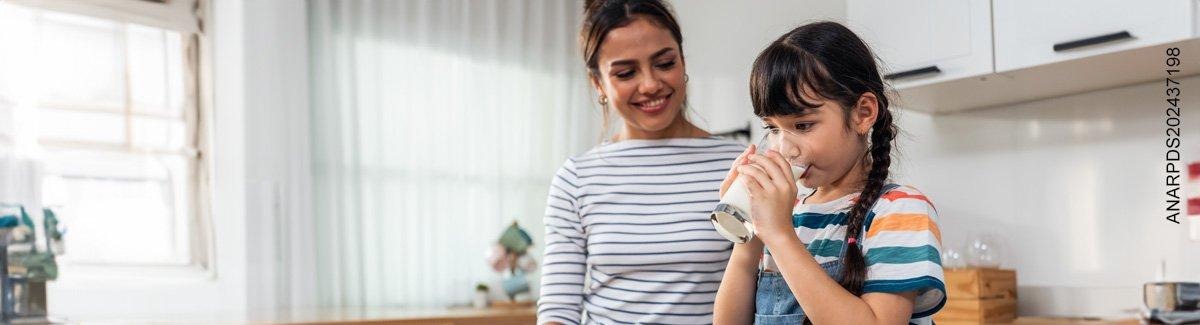 Niña toma de un vaso mientras su mamá la mira sonriendo.