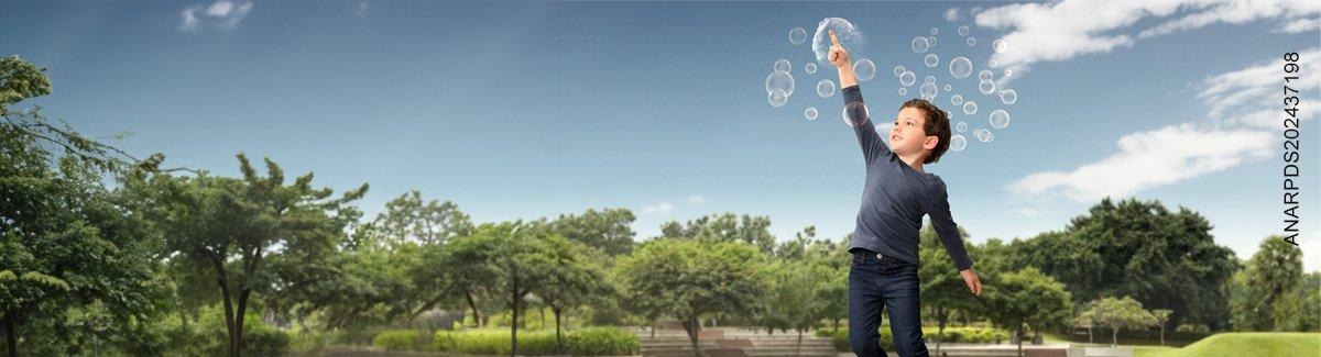 Niño, en un campo abierto con un bosque detrás de él, estalla una burbuja de jabón mientras sonríe.