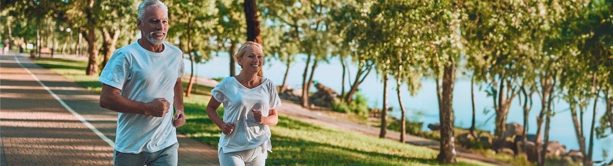 Pareja de adultos sonrientes trota por un parque al lado de un lago.