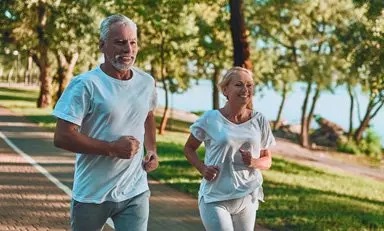 Pareja de adultos sonrientes trota por un parque al lado de un lago.