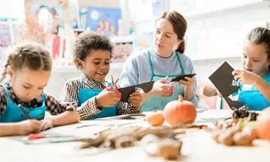 Profesora conversa con un niño mientras recortan una cartulina, dos niñas a su lado recortan y colorean.