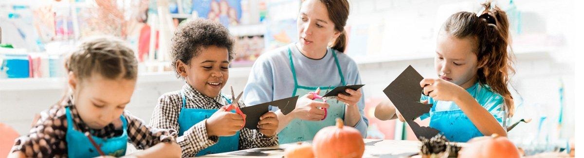 Profesora conversa con un niño mientras recortan una cartulina, dos niñas a su lado recortan y colorean.