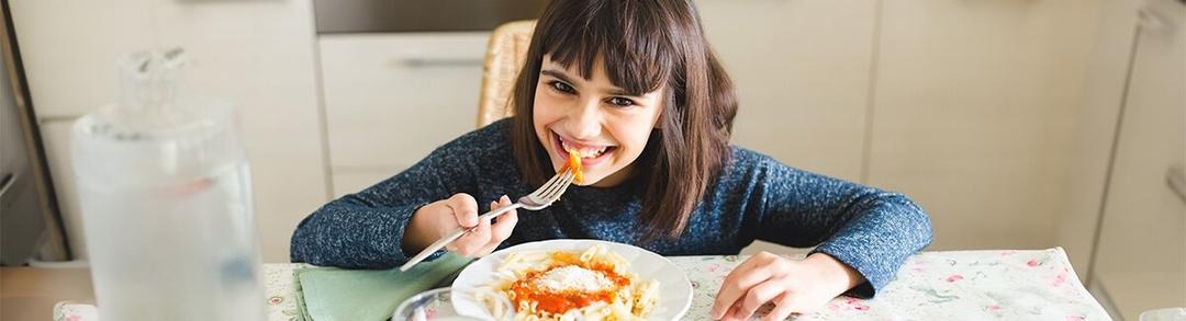 Dos niños sentados en una mesa en una cocina, cada uno bebiendo de un vaso. El niño a la izquierda tiene el cabello largo y sostiene el vaso con ambas manos