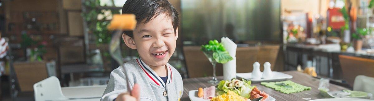Un niño sentado a una mesa con un plato de comida variada que incluye huevos revueltos, jamón, ensalada y una rodaja de zanahoria. El niño sostiene un tenedor con un trozo de comida. 