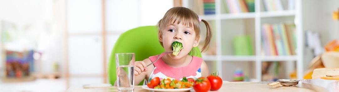 un niño sentado a la mesa con un plato de comida y un vaso de agua. El niño sostiene un tenedor y parece estar a punto de comer. El fondo incluye una silla verde y una estantería con varios objetos