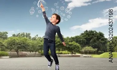Niño, en un campo abierto con un bosque detrás de él, estalla una burbuja de jabón mientras sonríe.